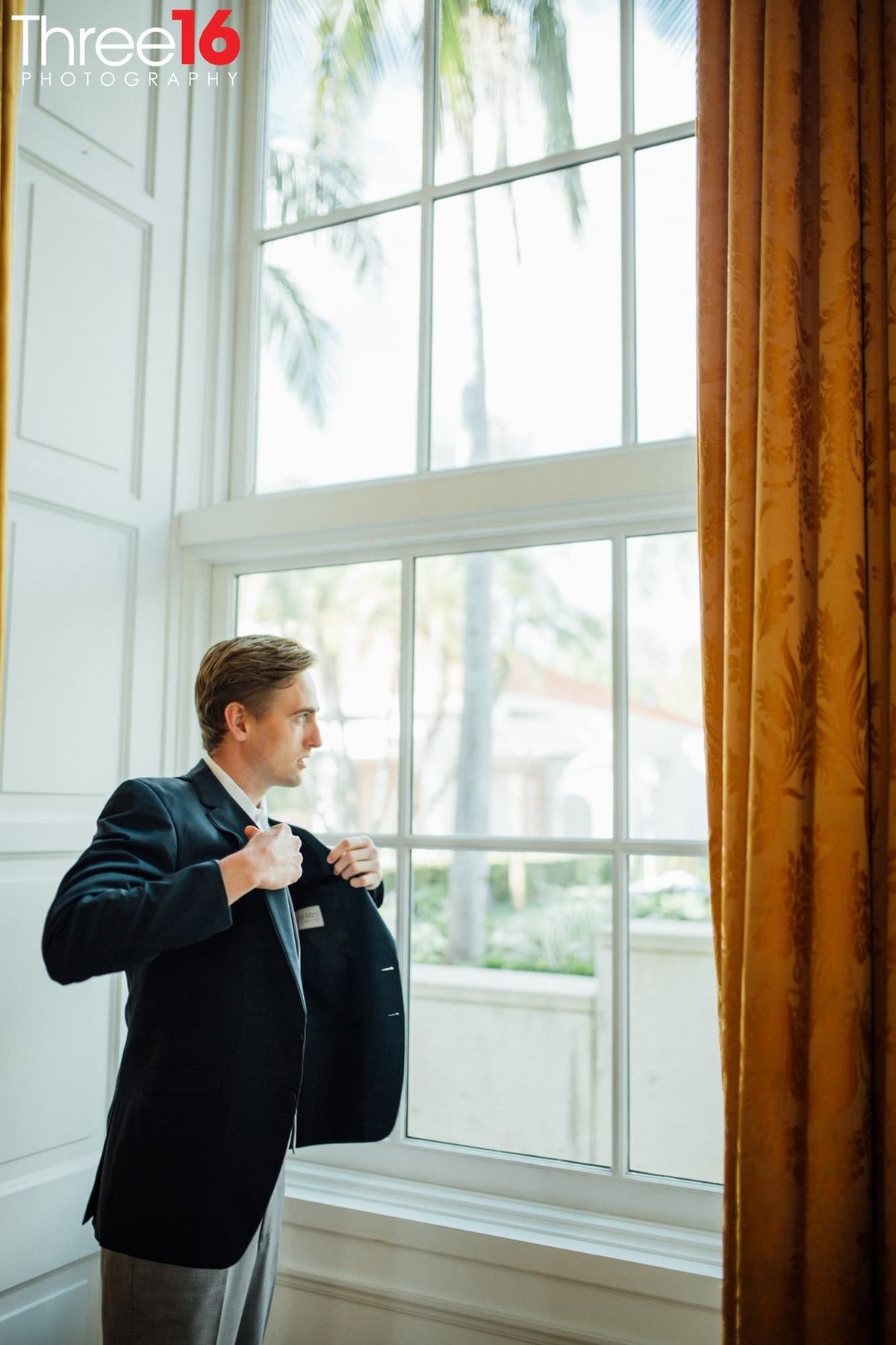 Groom looks out the window while putting on his coat before his wedding