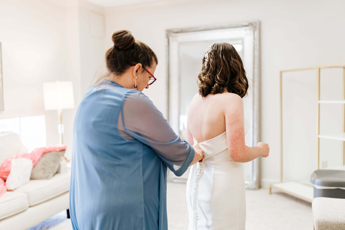 A woman in a blue robe helps a bride fasten the back of her white wedding dress in a bright, cozy room with a large mirror and neutral décor.