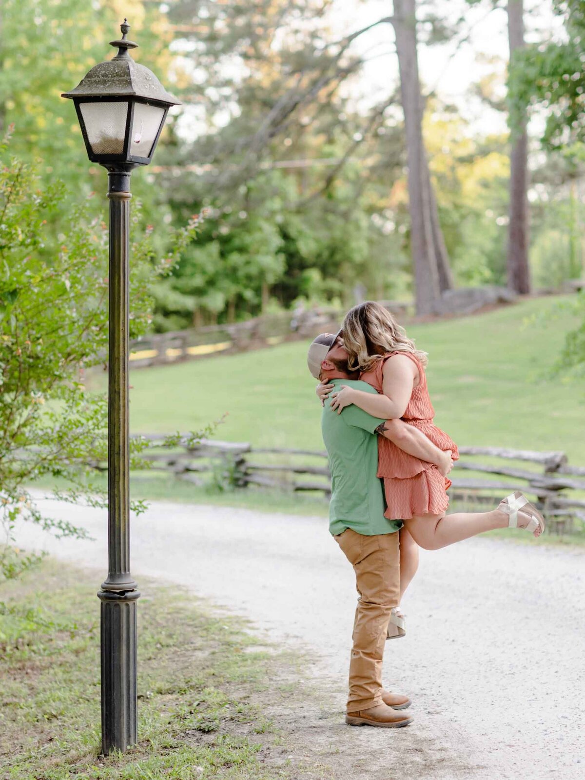 Man lifting his woman for a kiss on a gravel road with a lamp post in Fayetteville.