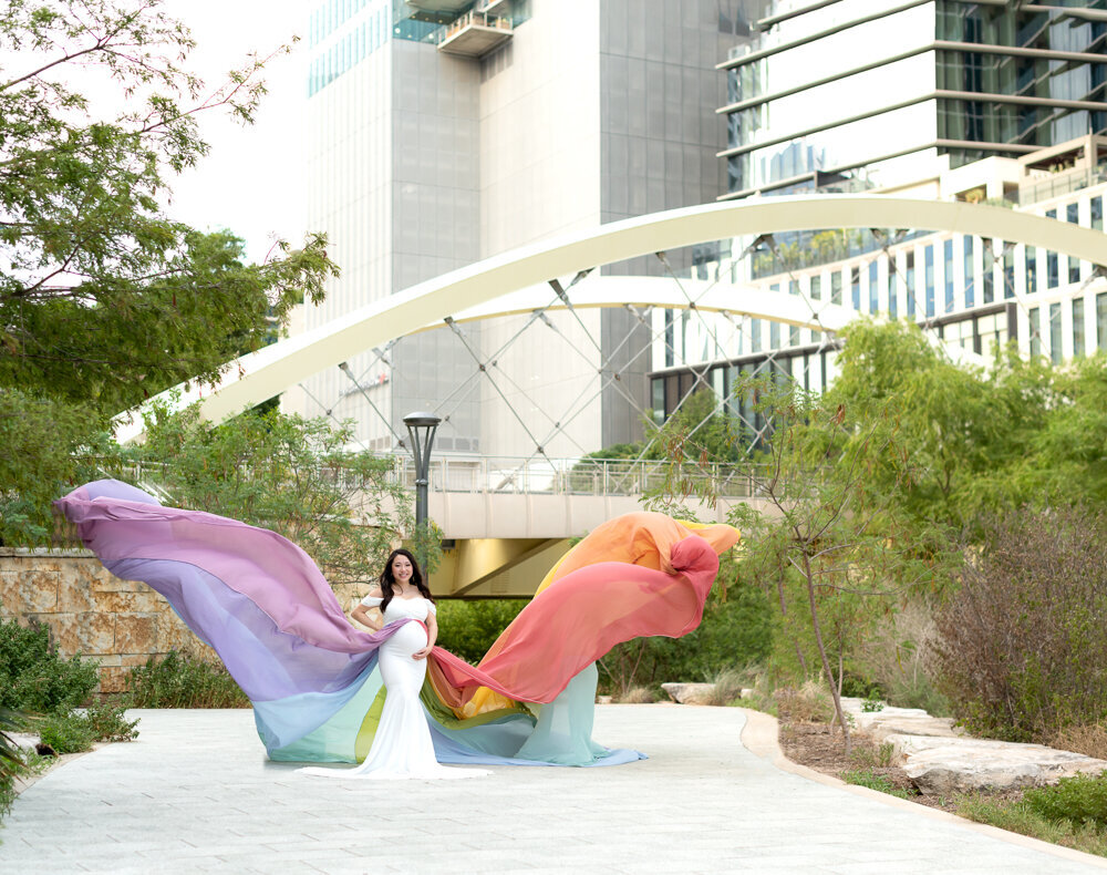 Elegant maternity portrait of a woman wearing a rainbow flowing gown in downtown Austin with a modern bridge backdrop.