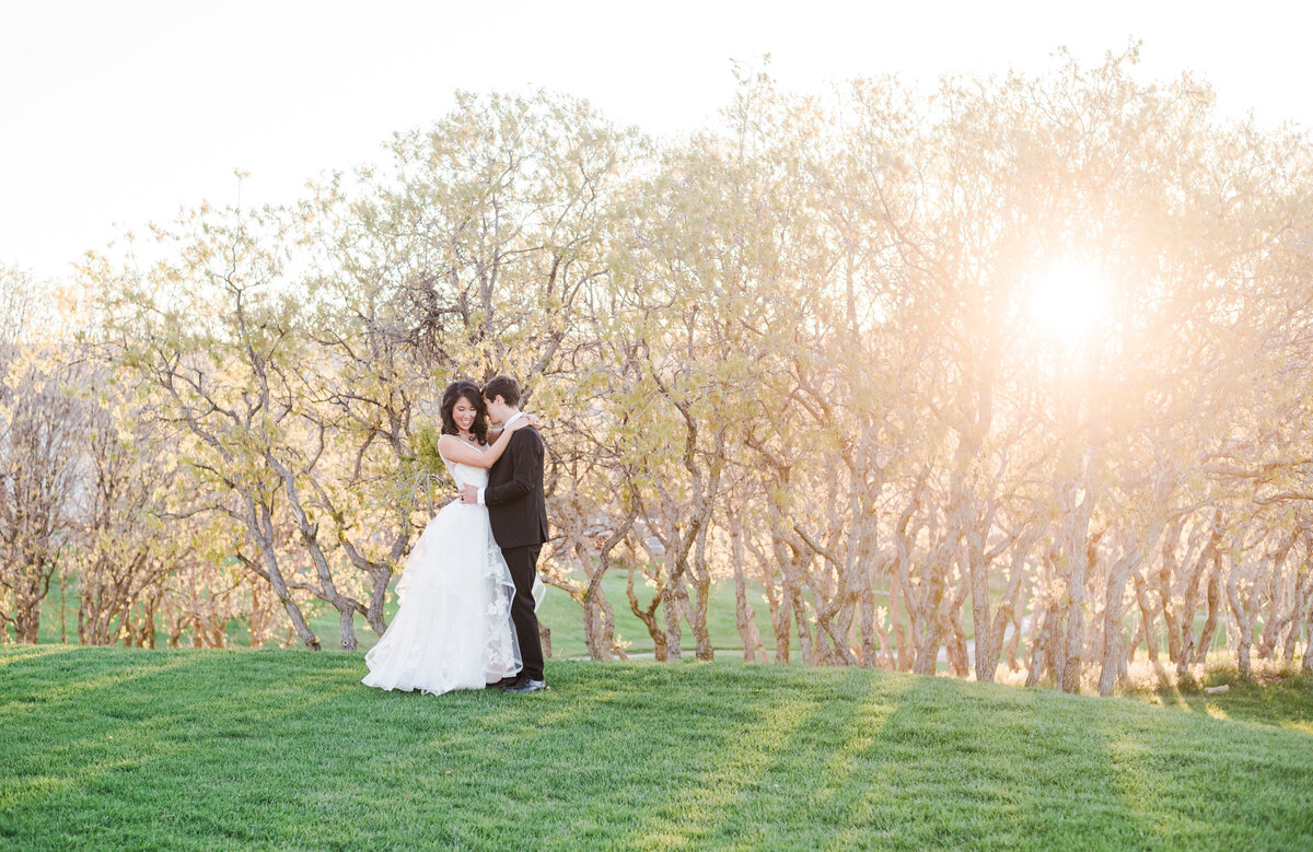 outdoor wedding portrait of bride adn groom embracing ina. green field with a row of trees in the distance letting the setting sun shine through them to the bride and groom captured by denver wedding photographer