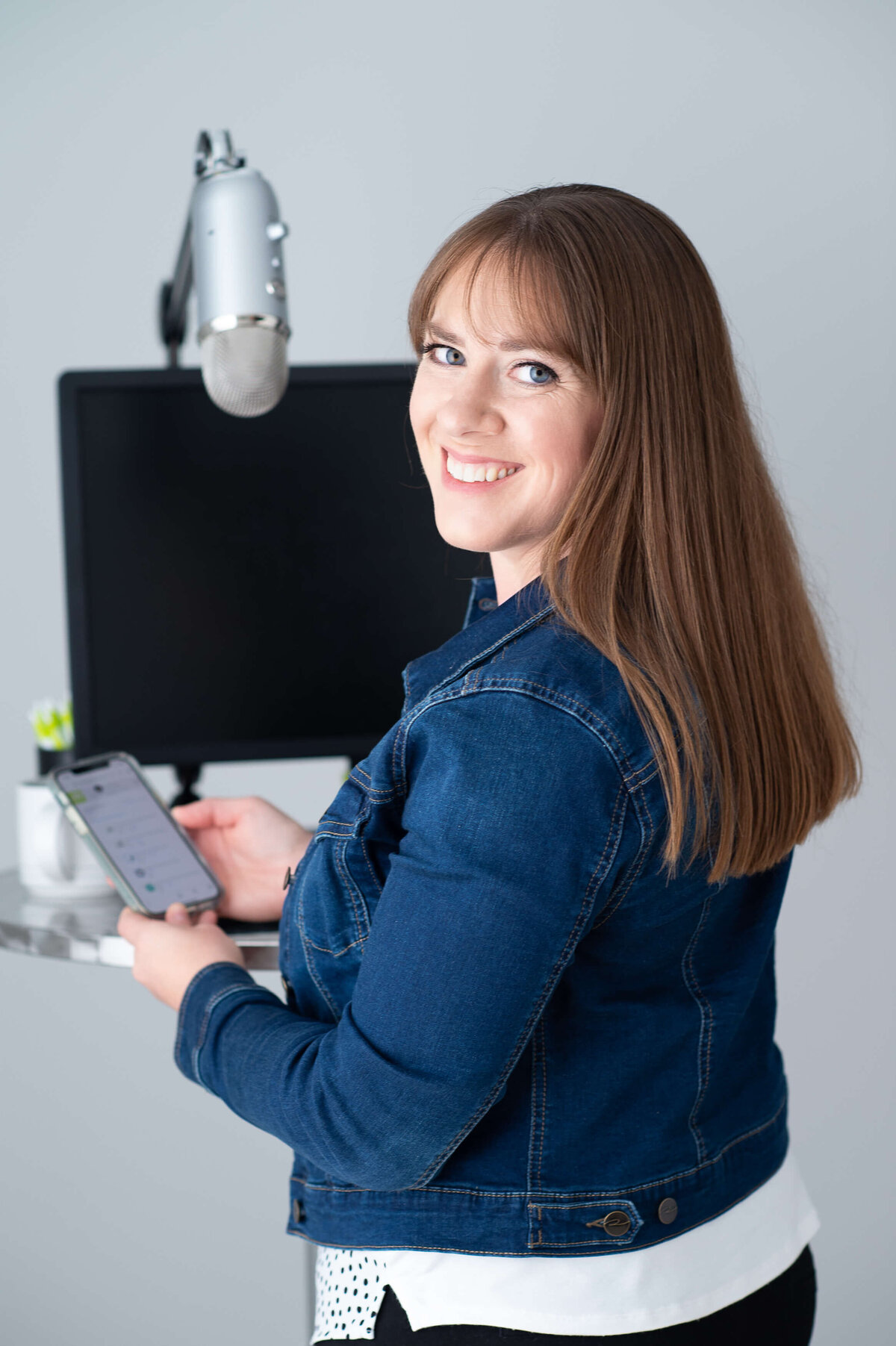 a digital marketing consultant wearing a jean jacket holding her cell phone in front of a computer and microphone.  Captured in studio by Ottawa Branding Photographer JEMMAN Photography Commercial