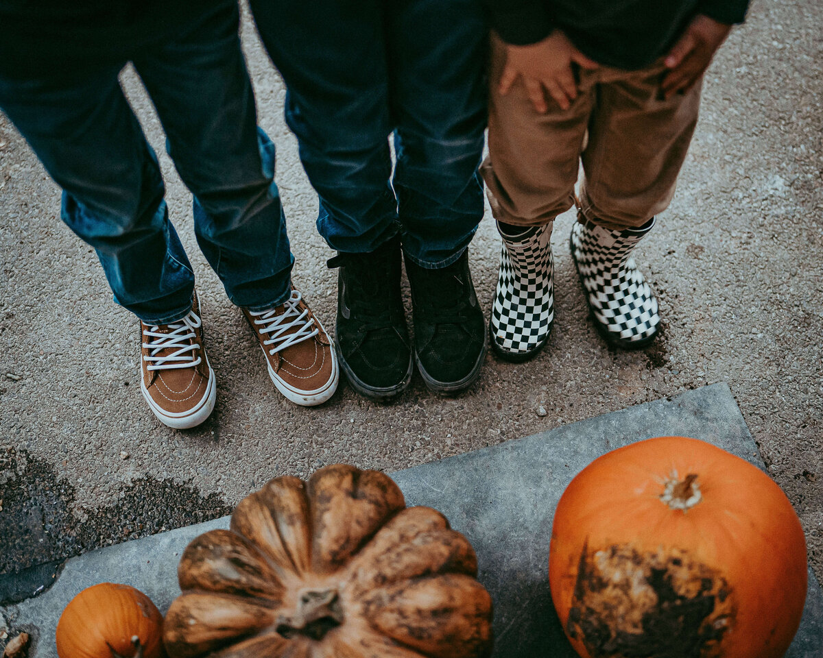 Three Brothers stand with her feet together next to the pumpkins that they grew on their family farm. This is a close-up picture with none of their faces just their feet and the pumpkins they grew..