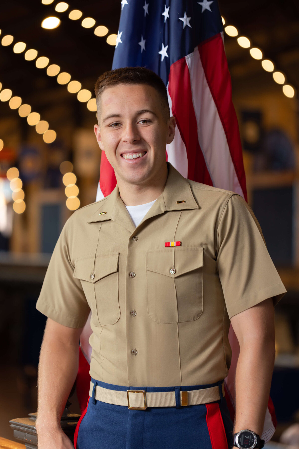 U.S. Marine Corps officer smiles with bokeh lights and flag in Naval Academy Dahlgren Hall.