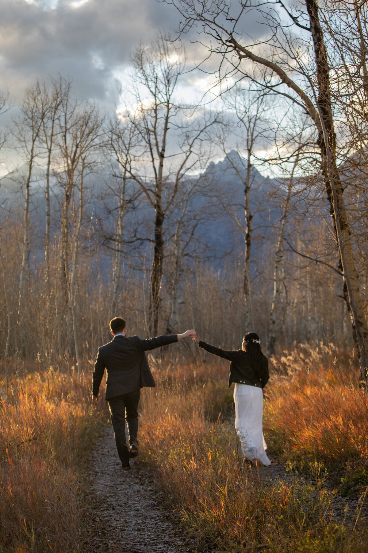 A bride and groom walk with their hands raised as they walk through a grassy dirt road.