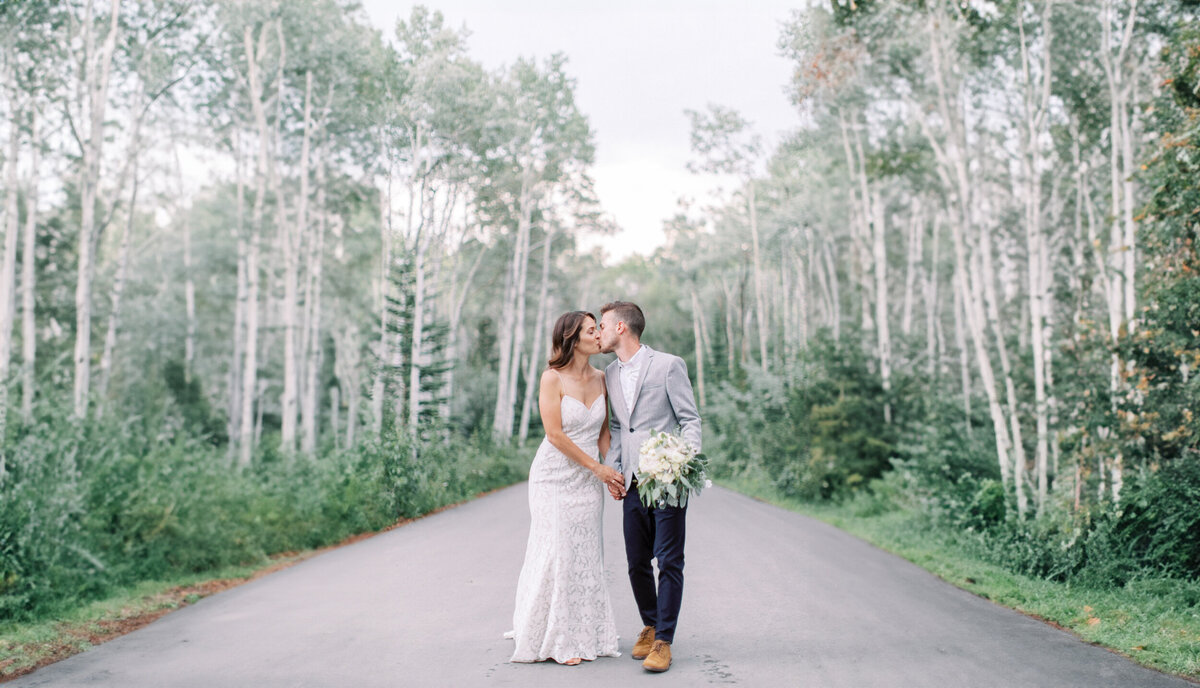 bride and groom walking and sharing a kiss on a road surrounded by birch trees at their Calgary wedding photographed by Calgary wedding photographers Heidrich Photography