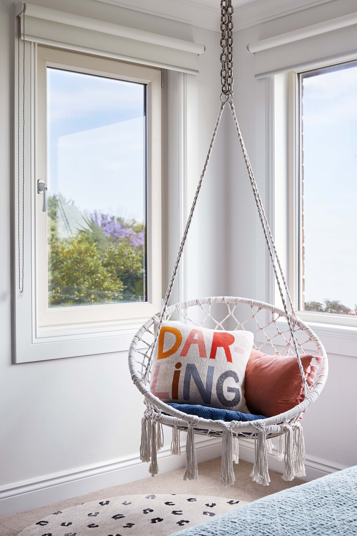 A corner of a room in a Williamstown residence featuring a white macrame hanging chair with a vibrant "DARLING" pillow and a rust-colored pillow, creating a playful and cozy reading nook.