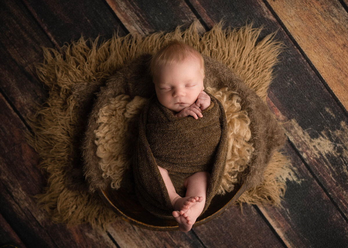 A baby sleeps in a woven basket in a brown swaddle on rustic wood floor