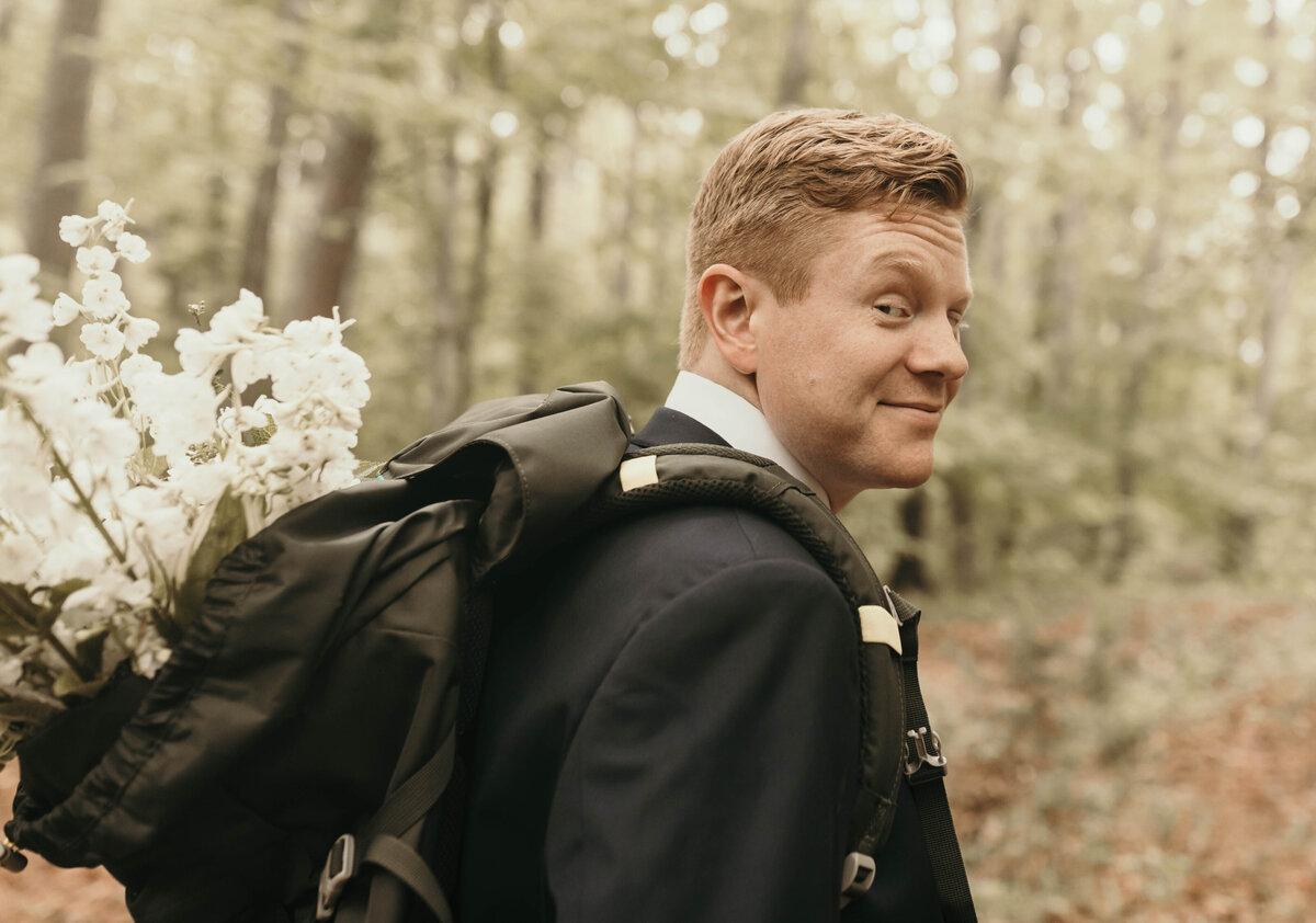 groom walking through woods with flowers in the backback