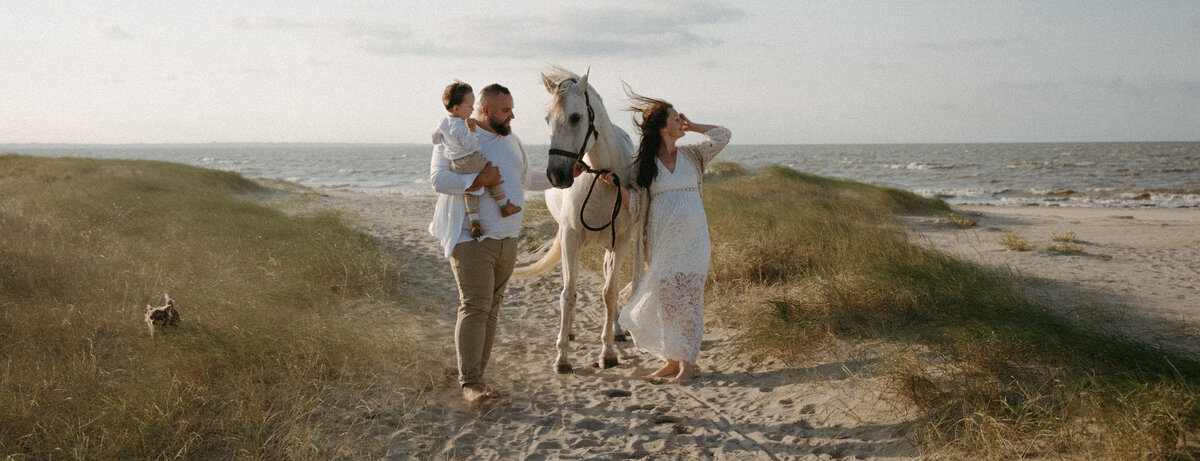 Papa portant son petit garçon dans les bras en compagnie de sa femme et de leur cheval blanc marchant dans les dunes face à la mer. Par Kamescop, photographe et vidéaste en Bretagne.