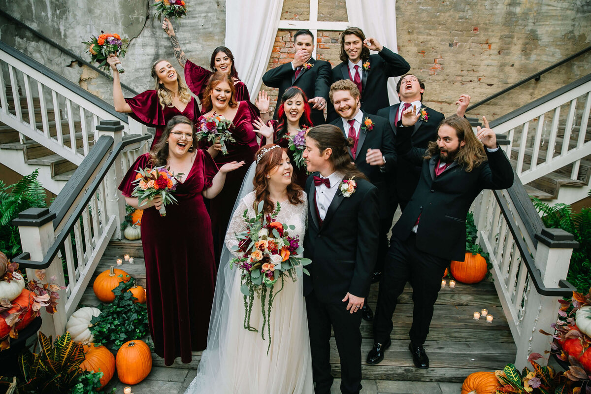 A bride and groom stand on stairs, smiling at each other, surrounded by a joyful wedding party in burgundy attire. The scene is decorated with pumpkins, flowers, and greenery. Everyone looks happy and celebratory.
