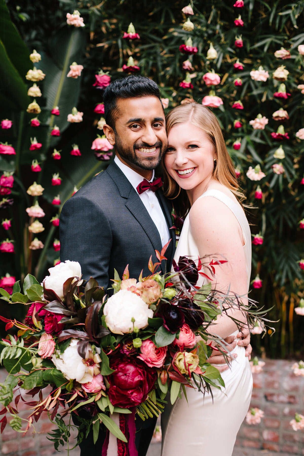 Sri-Lankan groom embraces his bride while they stand in front of a floral garland ceremony structure. Bride is holding a large burgundy bridal bouquet.