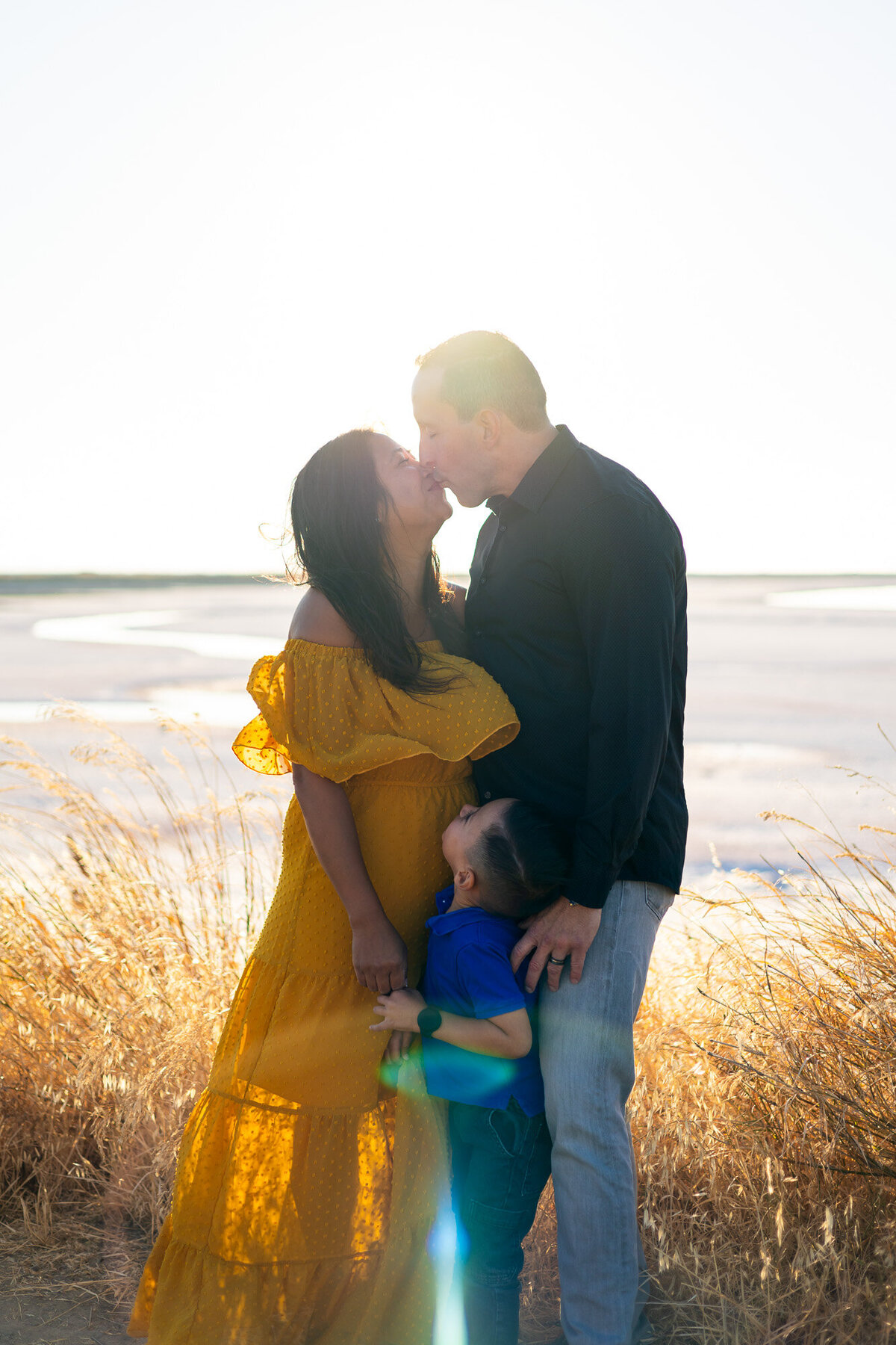 A candid family moment with parents sharing a kiss as their son looks up at them in golden hour light – Bay Area Family Portfolio - Ellobelle Photography