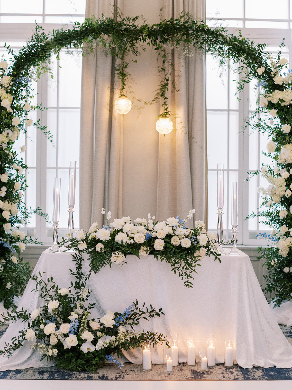 A beautifully decorated table with white flowers and greenery, set under an arch adorned with plants in front of large windows, perfect for a Wedding at Fairmont Palliser Calgary. Candles are placed on and around the table, creating an enchanting atmosphere.