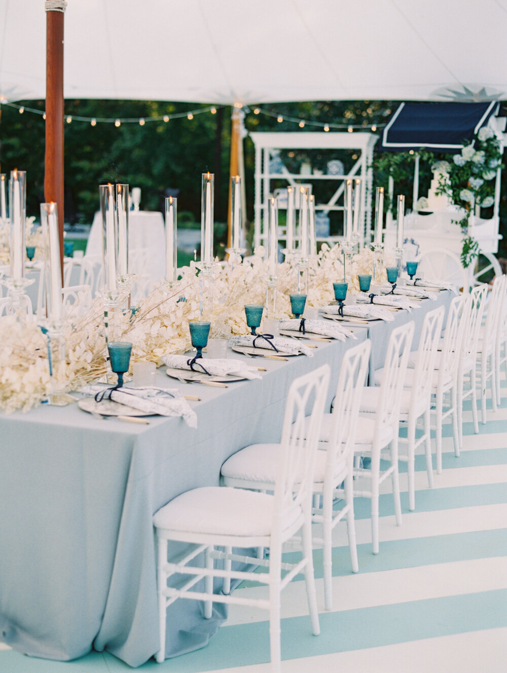 The stunning photo was taken at The Hamptons, New York, capturing a waterfront estate tented wedding through the lens of photographer Bonnie Sen, with East West Films as videographers. In the picturesque setting, a long, elegant dining table is elegantly arranged outdoors under a tent. Adorned with a light blue tablecloth, white chairs, and blue glassware, the table exudes sophisticated charm. Tall, slender candleholders and a captivating white floral centerpiece grace the length of the table, adding a touch of romance to the scene. Lush foliage and twinkling string lights beautifully frame the celebratory ambiance in the background.