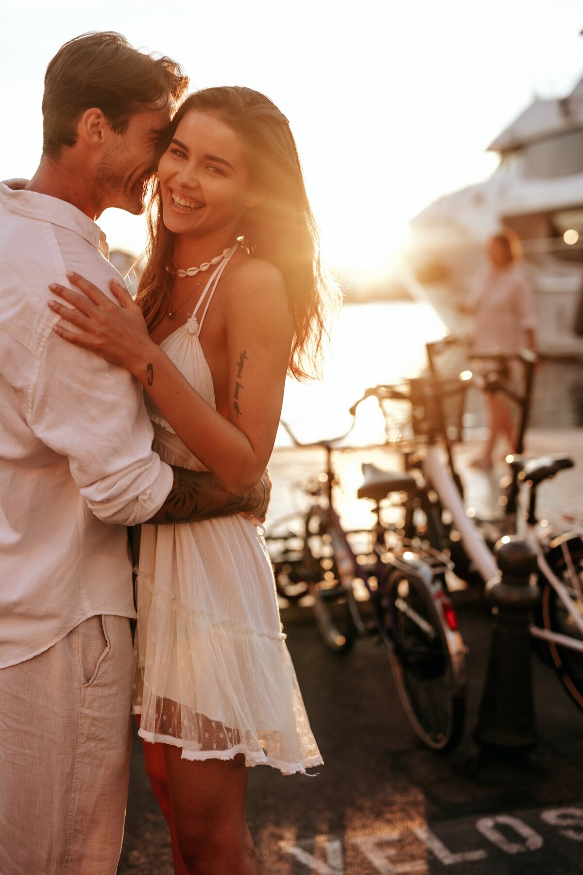 Couple looking at the camera and smiling while embracing each other with the sun coming down behind them and a yacht in the background.