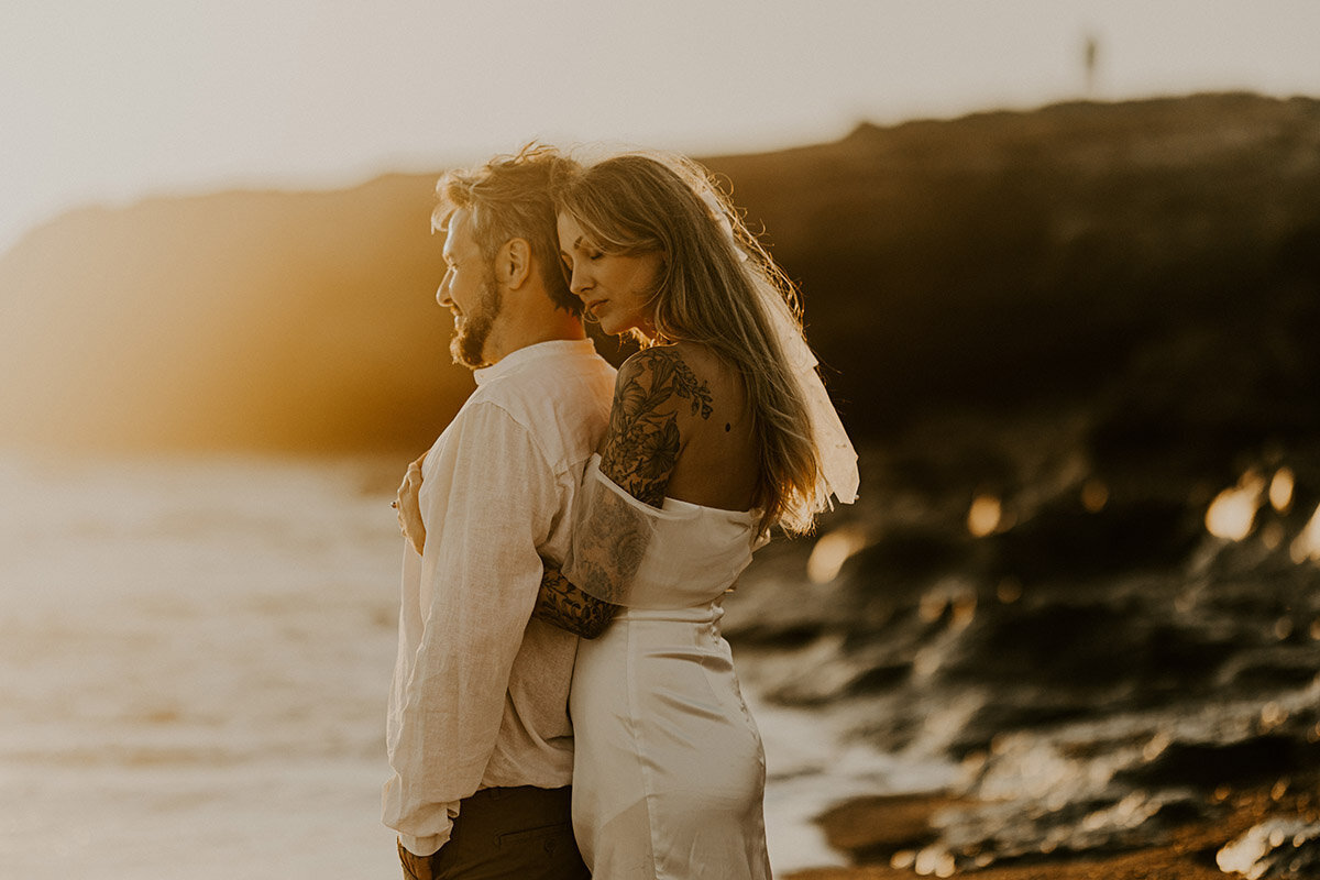 Femme serrant son compagnon de dos sur une plage au coucher du soleil. Moment capturé par Laura, photographe couple en Vendée.