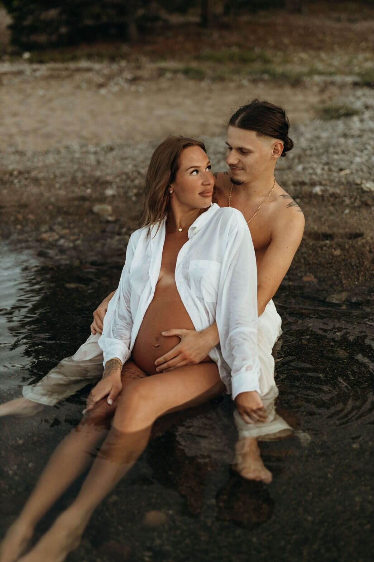 Gravid kvinna sittandes framför sin partner i vattnet och blir omhållen under en gravidfotografering i Stockholm