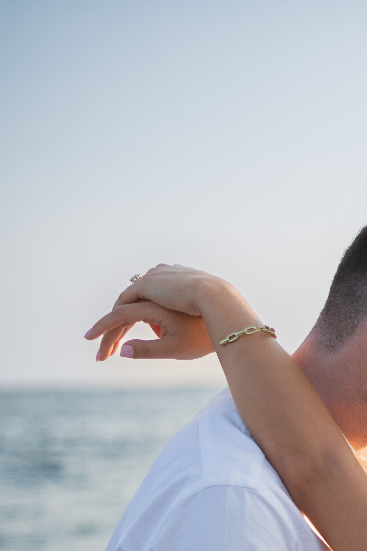 Collins Beach Newport RI | Kelsey Sheehan Photography Timeless Rhode Island Weddings | Close-up of a couple embracing by the sea. A woman's hand rests gently on a man's shoulder, wearing a gold bracelet, conveying intimacy and tranquility.