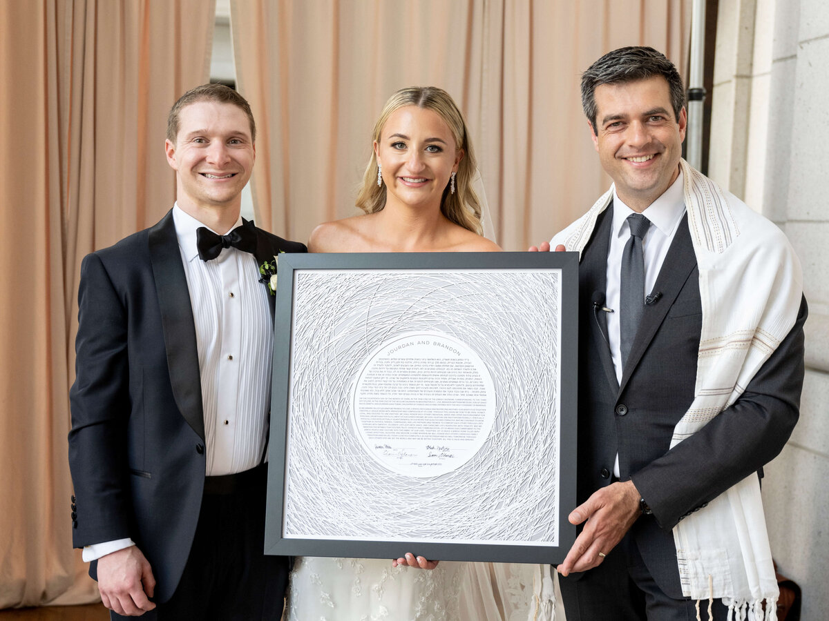 A bride, groom, and rabbi smile, holding a framed document with intricate designs. The setting appears to be indoors with soft drapes in the background.