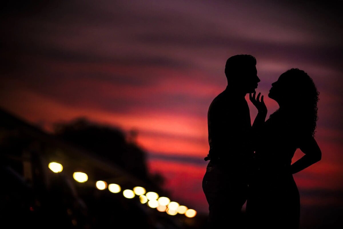 Silhouette of a couple against a vivid crimson sky after sunset, with lights twinkling in the background