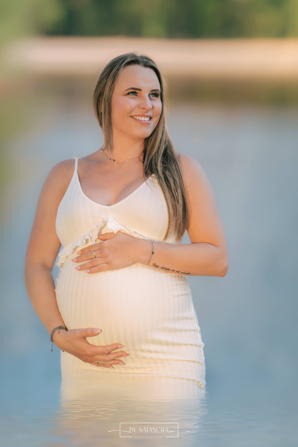 Vrouw kijkt glimlachend opzij, houd haar zwangere buik vast met hand erboven en eronder in een witte jurk met blauw helder water op de achtergrond