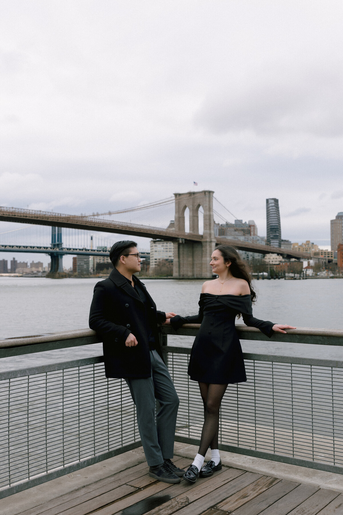 Romantic Brooklyn Bridge Park engagement photo of couple in black outfits smiling at each other with East River and Brooklyn Bridge in the background