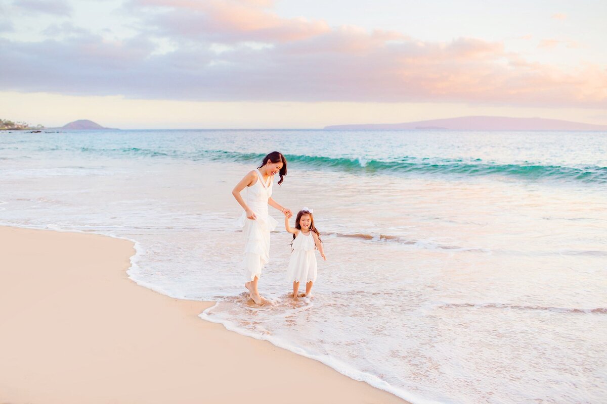 Woman with long black hair wears a white dress and holds her daughter's hand during their family session. Daughter is also wearing a white dress and looks off camera at the ocean