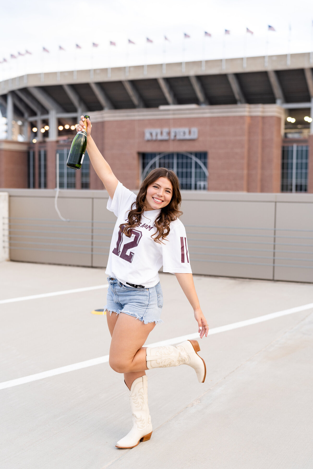 Texas A&M senior girl holding up champagne and celebrating while wearing white Aggie jersey on top of rooftop parking garage with Kyle Field in background