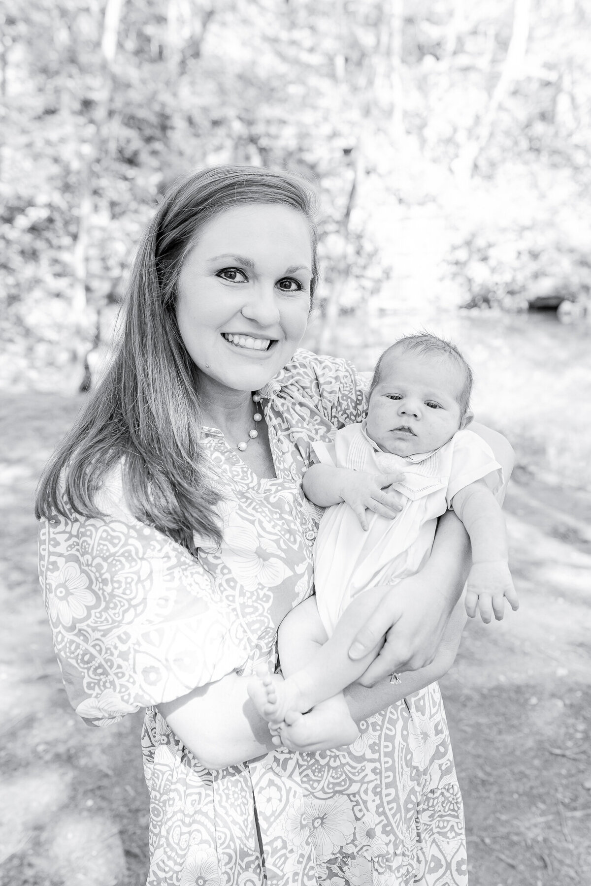 Mother holding newborn in a serene outdoor setting – captured by a Birmingham newborn photographer. A glowing mother beams with joy as she cradles her sweet baby, dressed in a classic heirloom outfit, surrounded by the peaceful beauty of nature.
