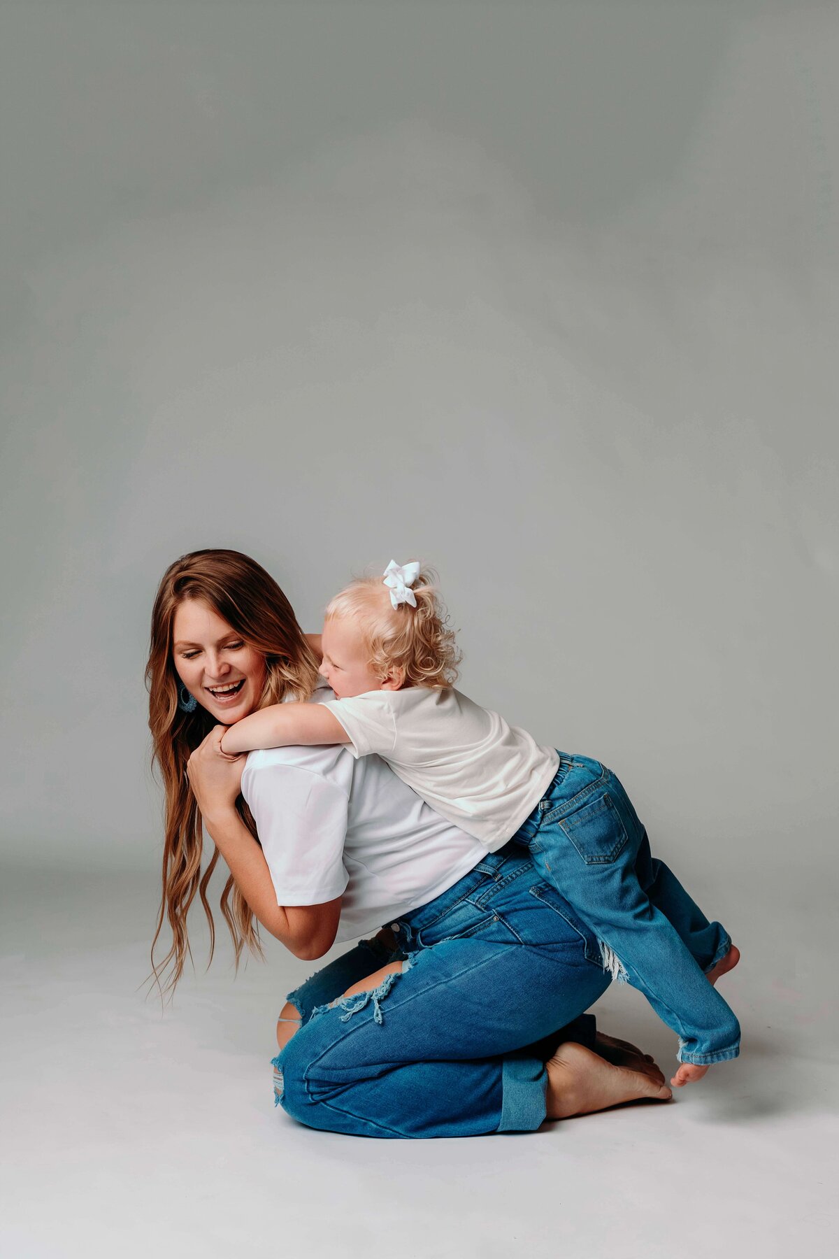 A young mom plays happily with her toddler as she sits on her knees and lets the child climn on her back. The relaxed portrait shows their unique bond, while they sport matching jeans and shirts in the studio of Mobile, AL motherhood photographer