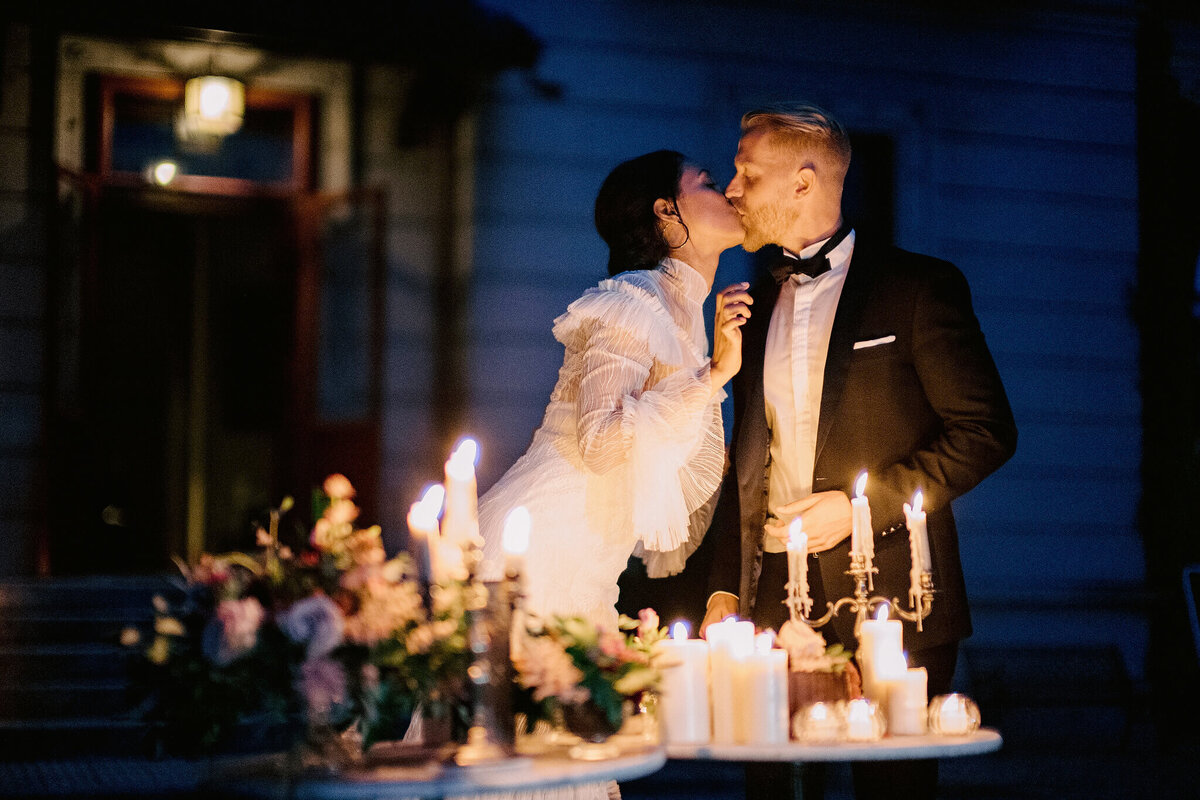 The bride and groom are standing while kissing with a table filled with candles and flowers, and a villa on the background.