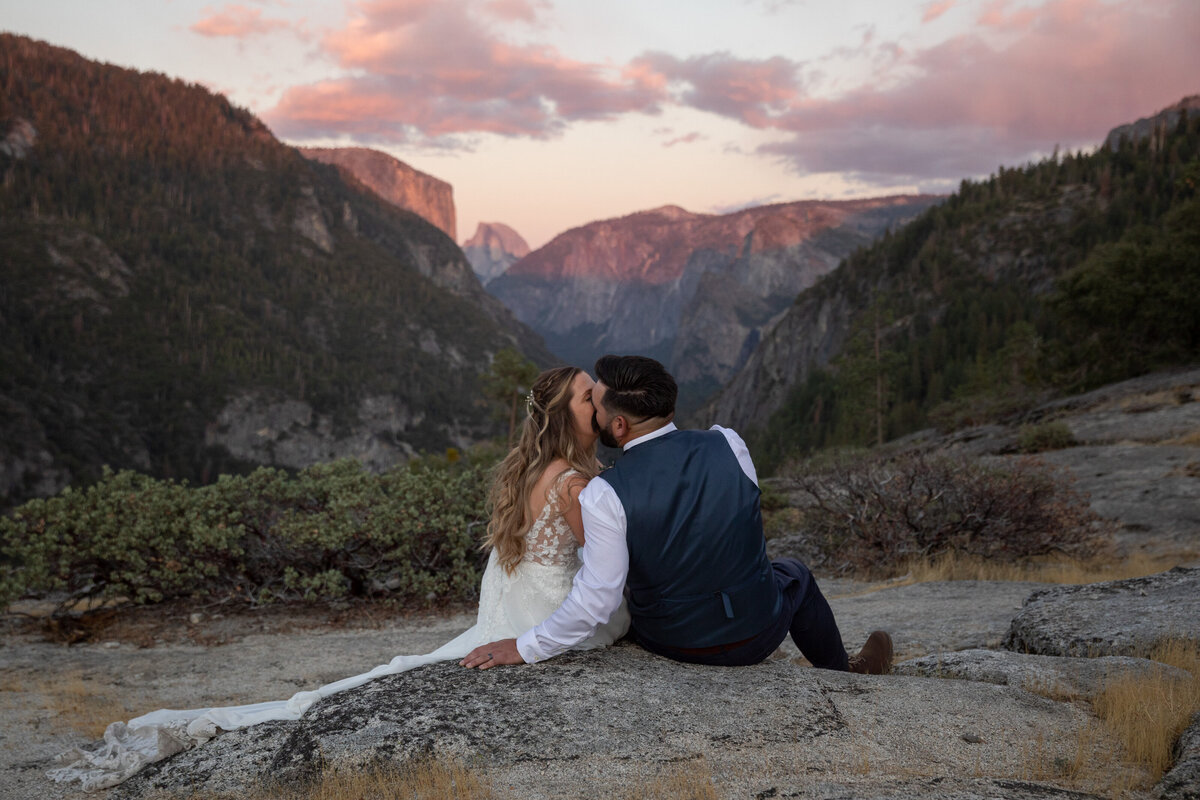 Bride and groom sit on a rock ledge at sunset, sharing a kiss with yosemite's cliffs and valley 