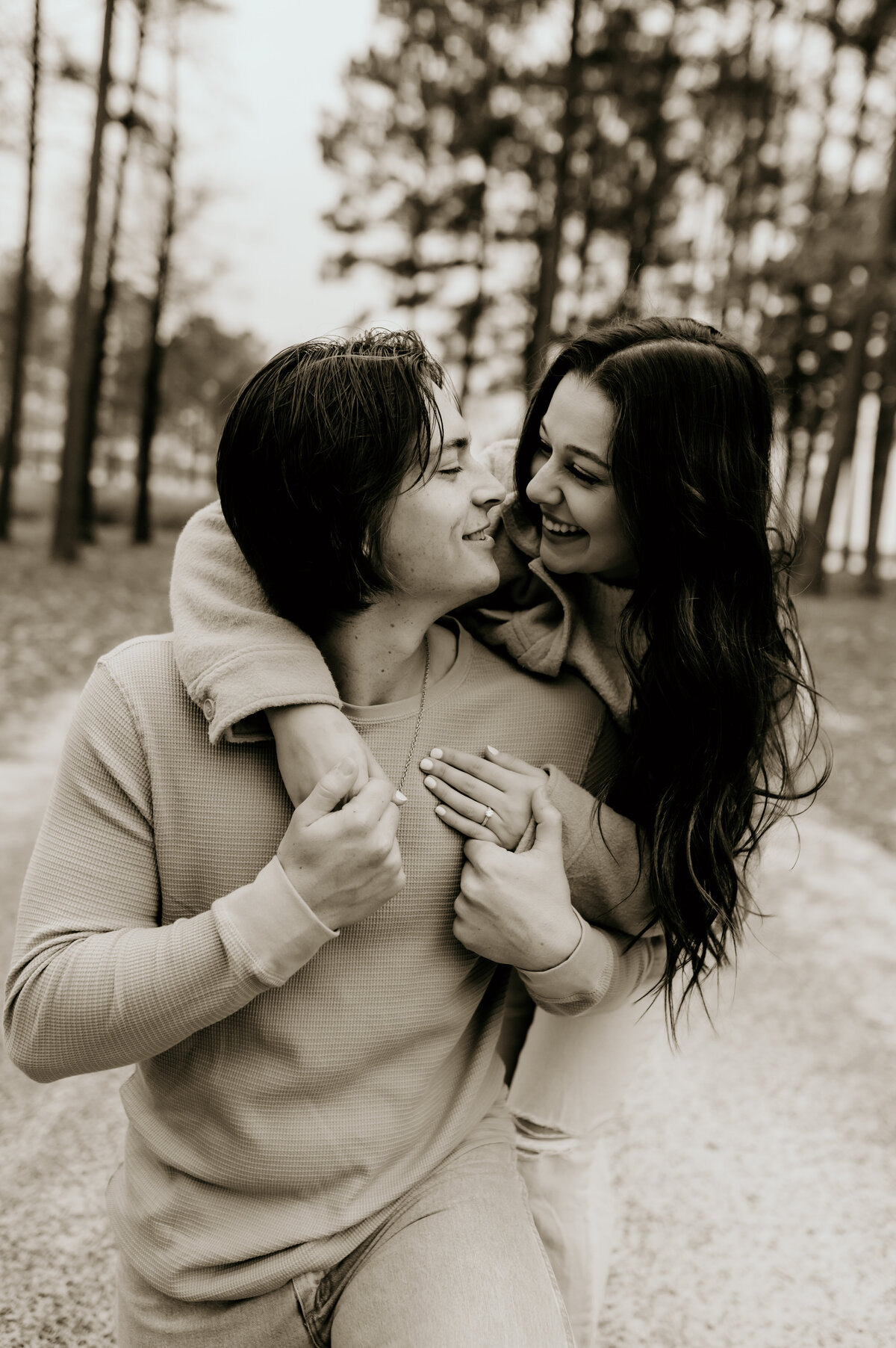 black and white engagement photography with woman standing behind a man and leaning over his shoulder with her arms wrapped around him and smiling down at him