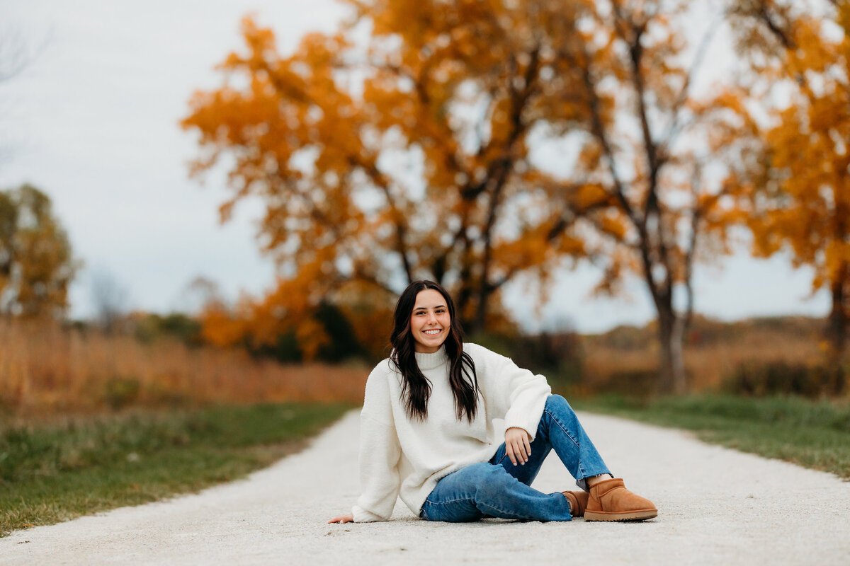A brunette sits on a gravel road with a knee up in the fall.