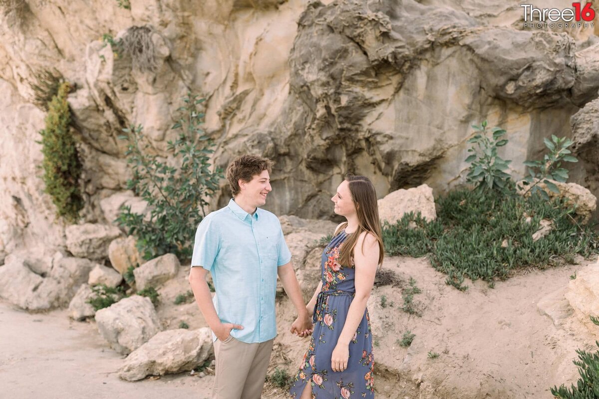 Engaged couple hold hands in front of a cliff at Corona del Mar State Beach