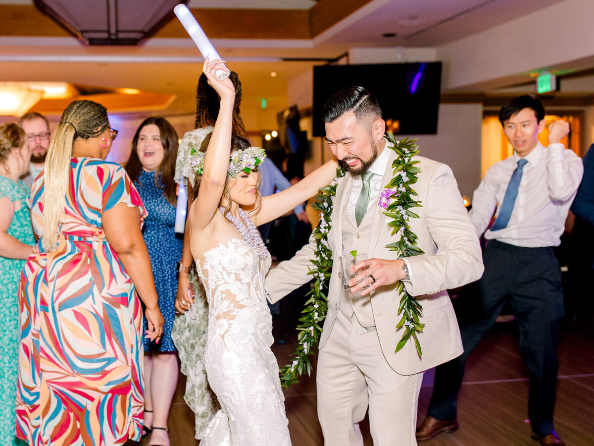 Bride and groom dance joyfully at a wedding, surrounded by guests in vibrant attire. The bride raises her arm, exuding happiness and celebration.