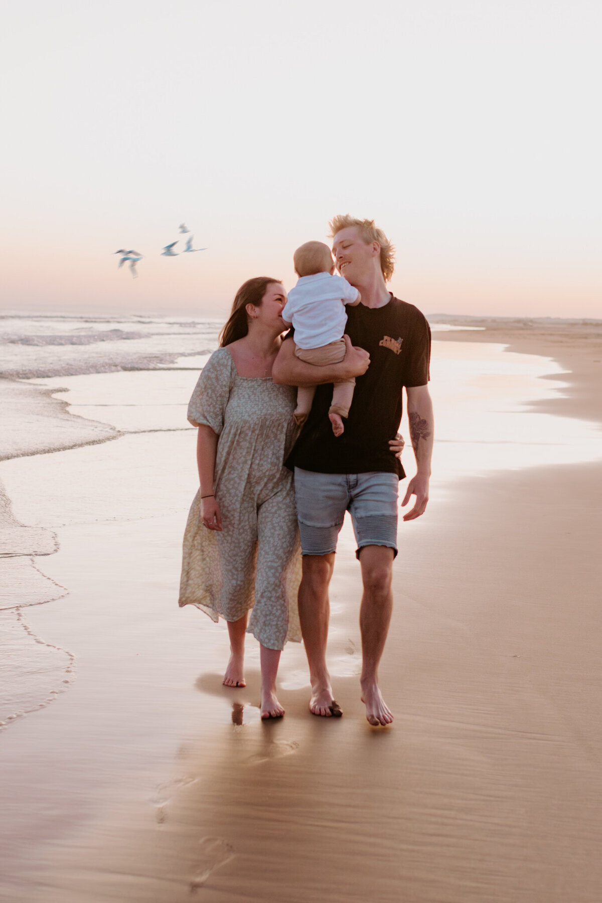 Young family walking barefoot along the beach at sunset, with parents smiling and holding their baby, captured during a relaxed family photo session by the ocean.