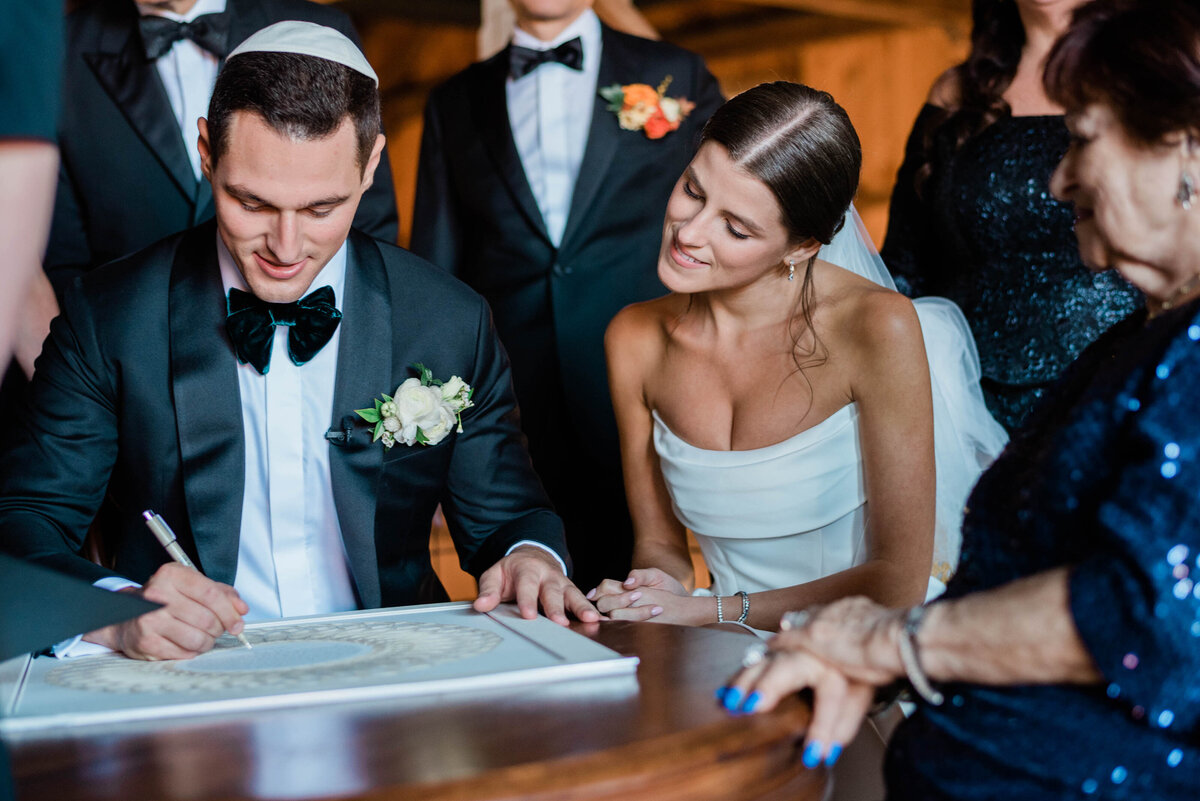 A groom, wearing a tuxedo, signs the ketubah at a Jewish wedding ceremony while the bride in a white gown watches