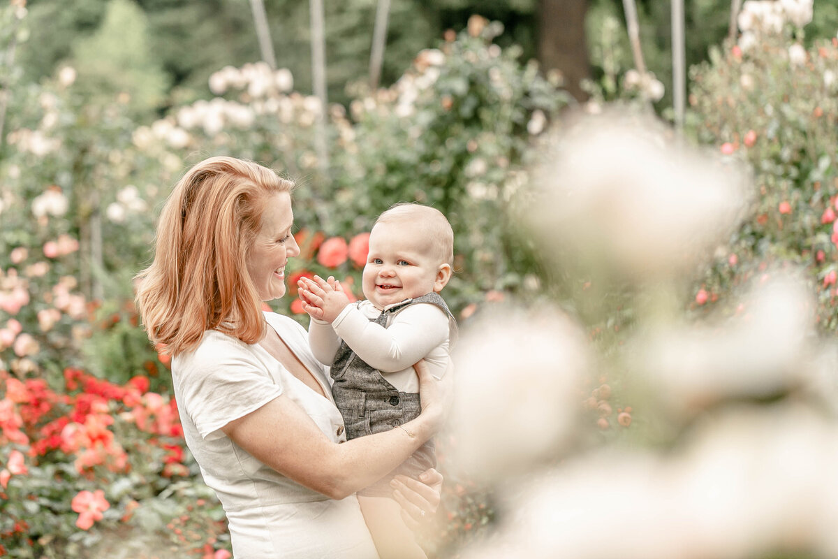 Mama in white dress holding one year old baby in light neutral tones. Mama is smiling at baby and baby is looking towards the camera and clapping. They are in the International Rose Test Gardens in Portland, Oregon.