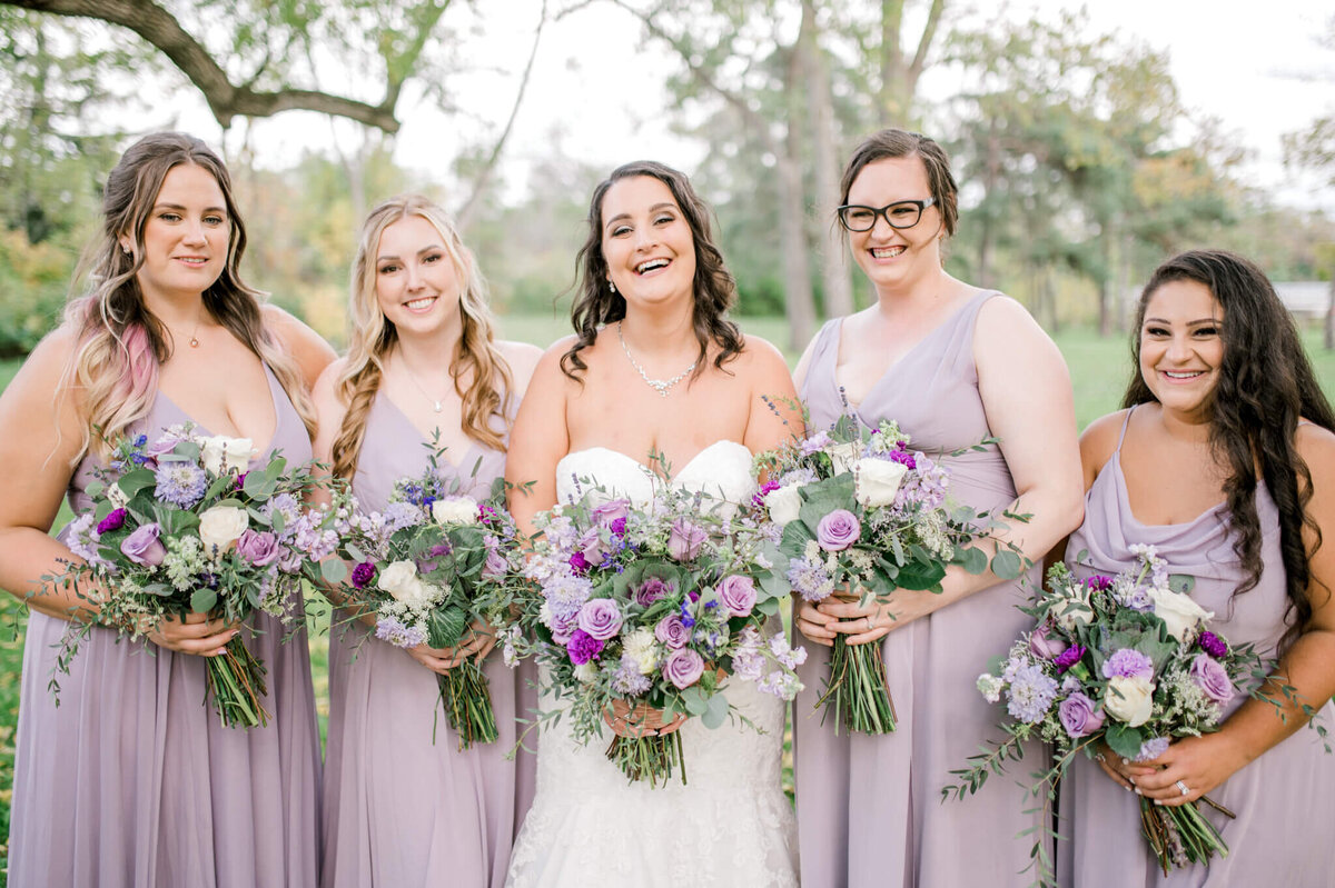 Bridesmaids in light purple dresses gathered around bride holding their bouquets at belly height  smiling  at camera.