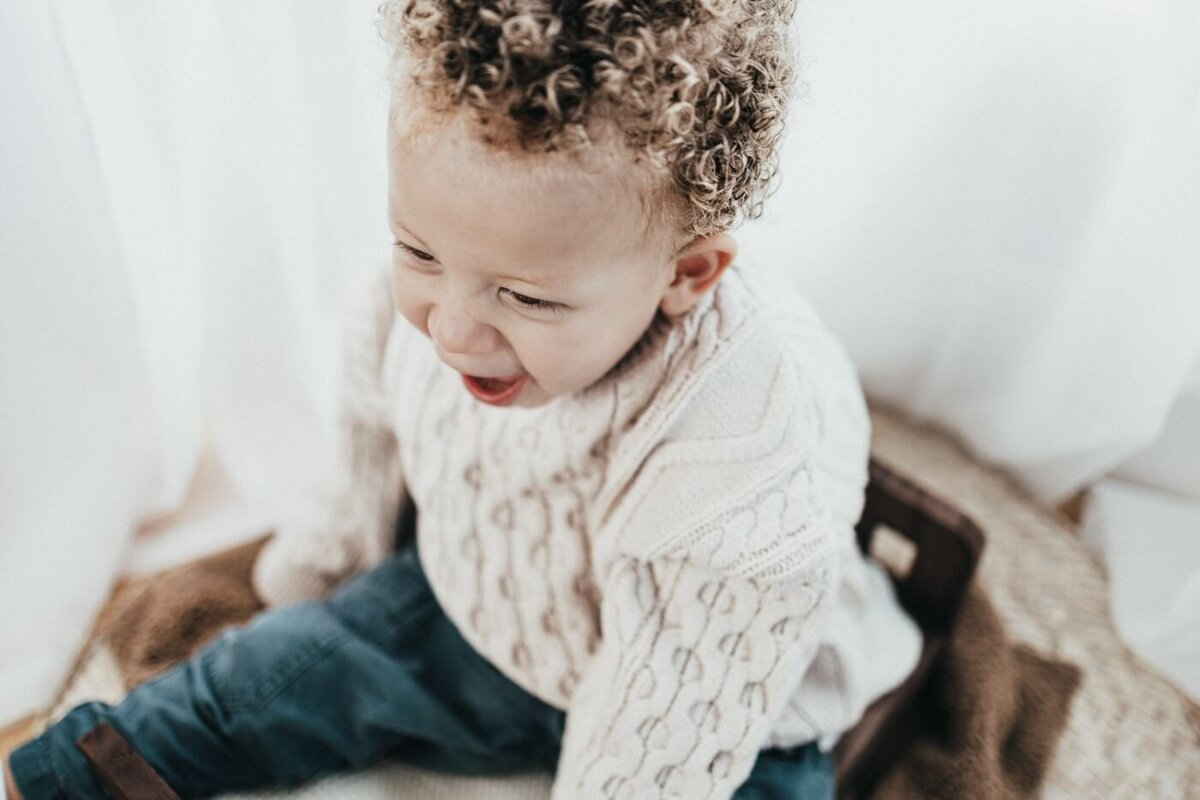 A joyful toddler with tight curly blonde hair and a big open-mouthed smile looks downwards in a bright setting