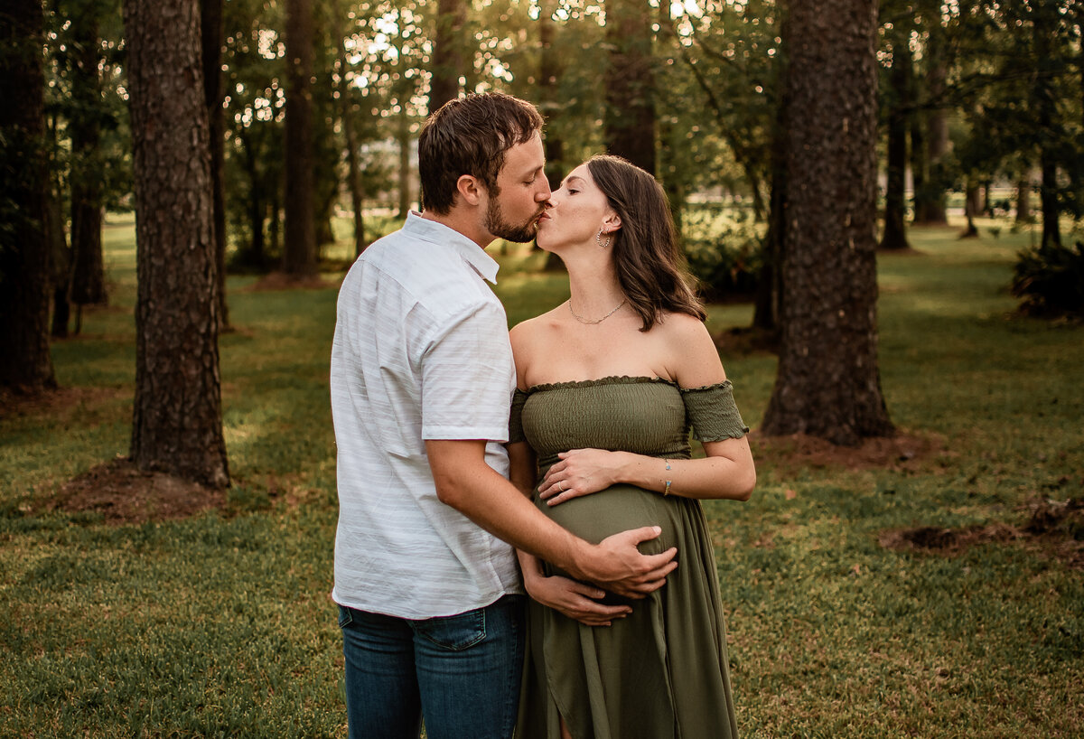 A mom and dad to be embrace and kiss in a field of pine trees.