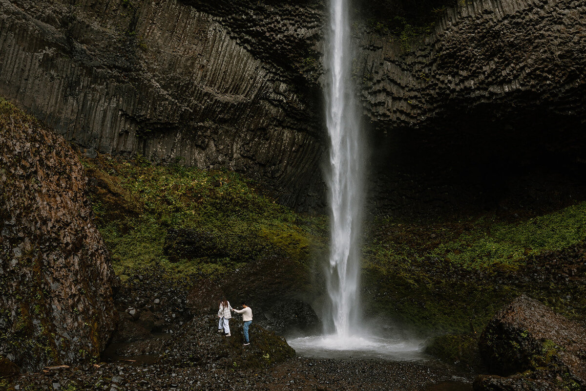 Jamie and Jordan’s adventurous waterfall engagement session in Oregon, walking hand in hand near cascading falls.