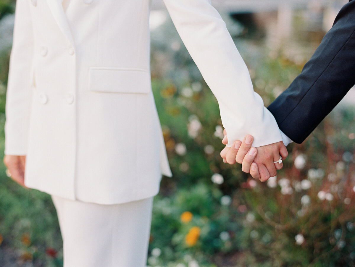 couple holding hands close up of round cut diamond engagement ring. bride is wearing white pant suit.