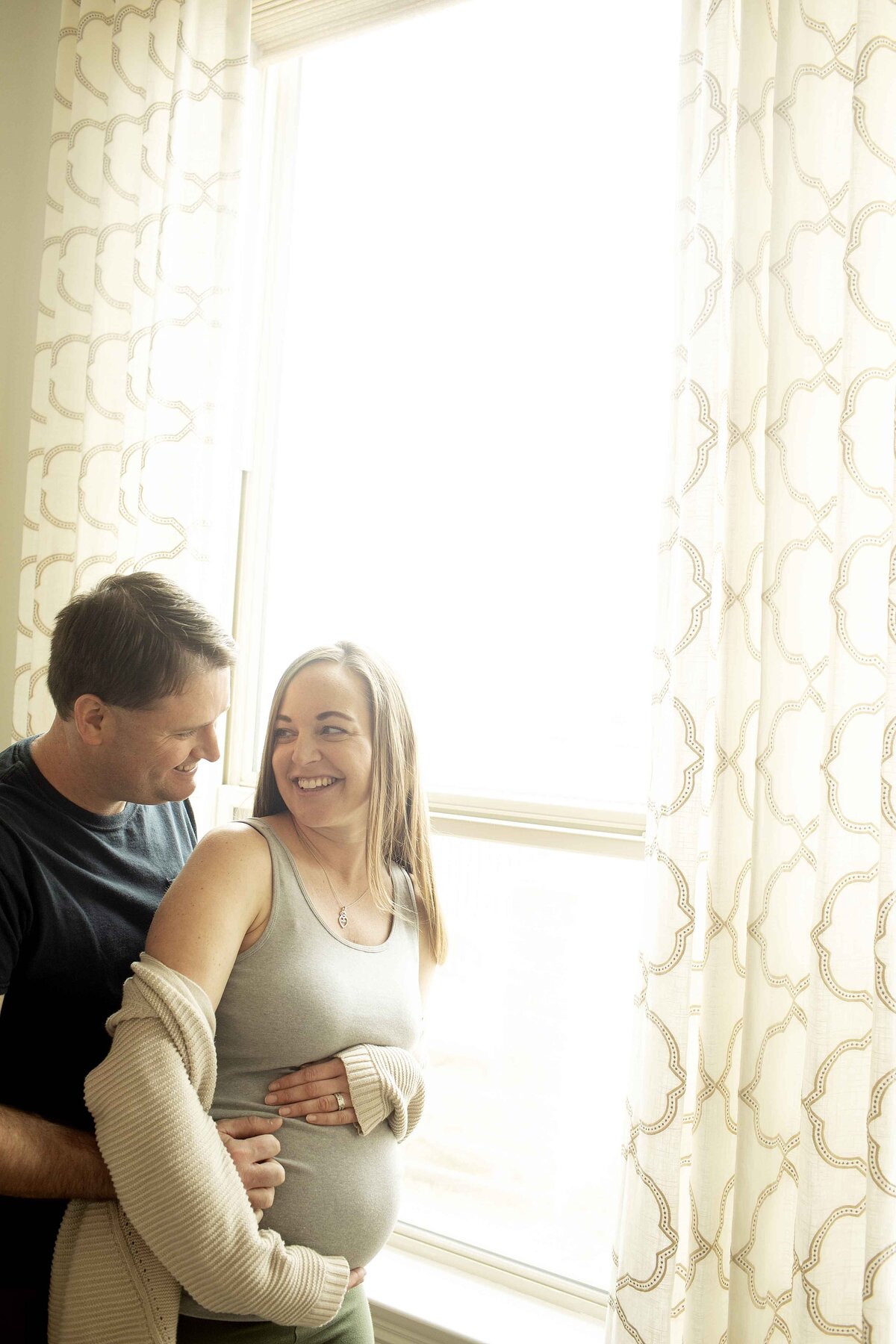 A beautiful maternity portrait of mom and dad in front of a large window, with light filtering through as they share a tender embrace, mom cradling her belly and dad gently kissing her forehead, captured by Chunky Monkey Photography.