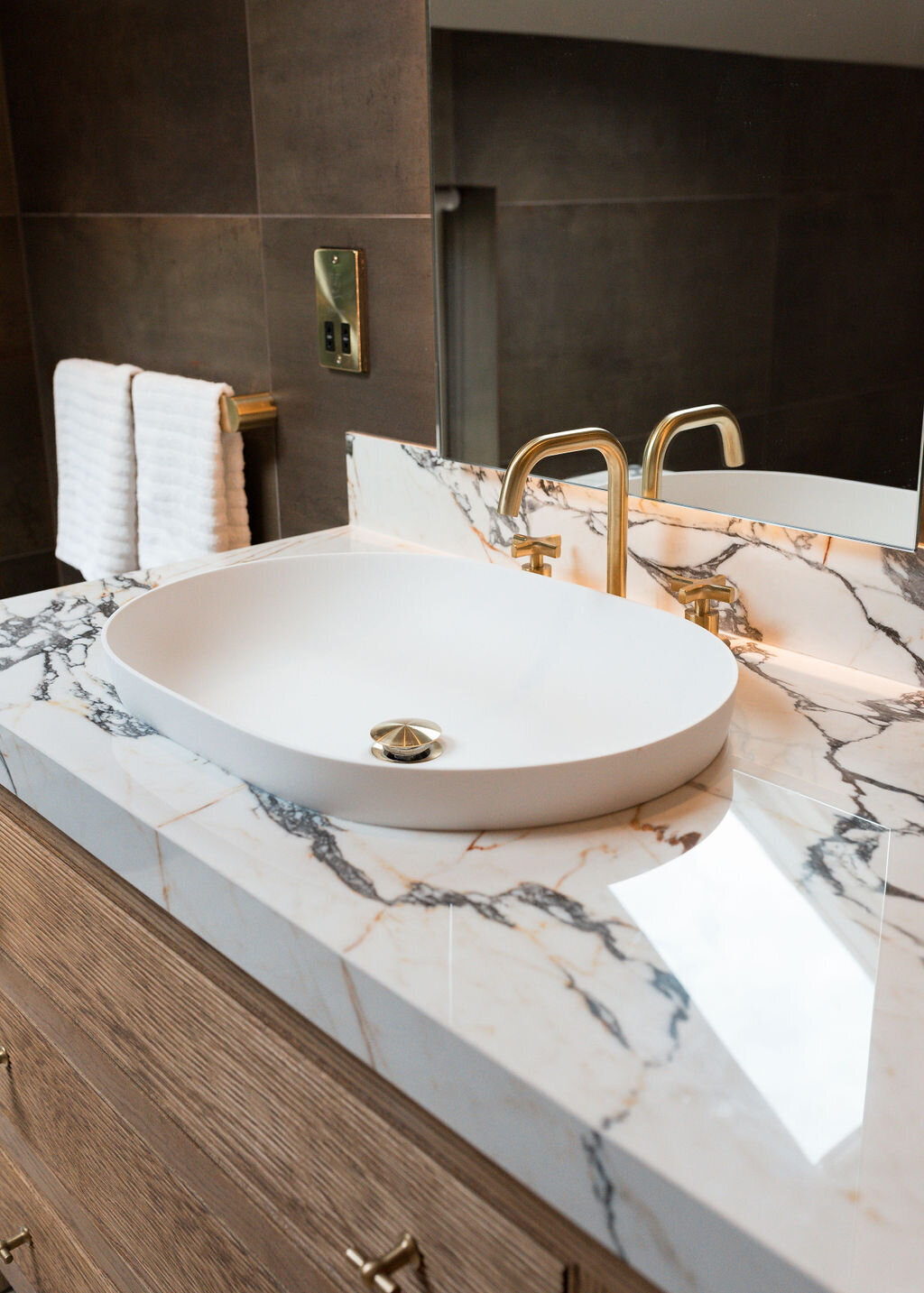 A modern bathroom with a double sink vanity with marble countertops. The sinks feature a white bowl and shiny gold hardware.