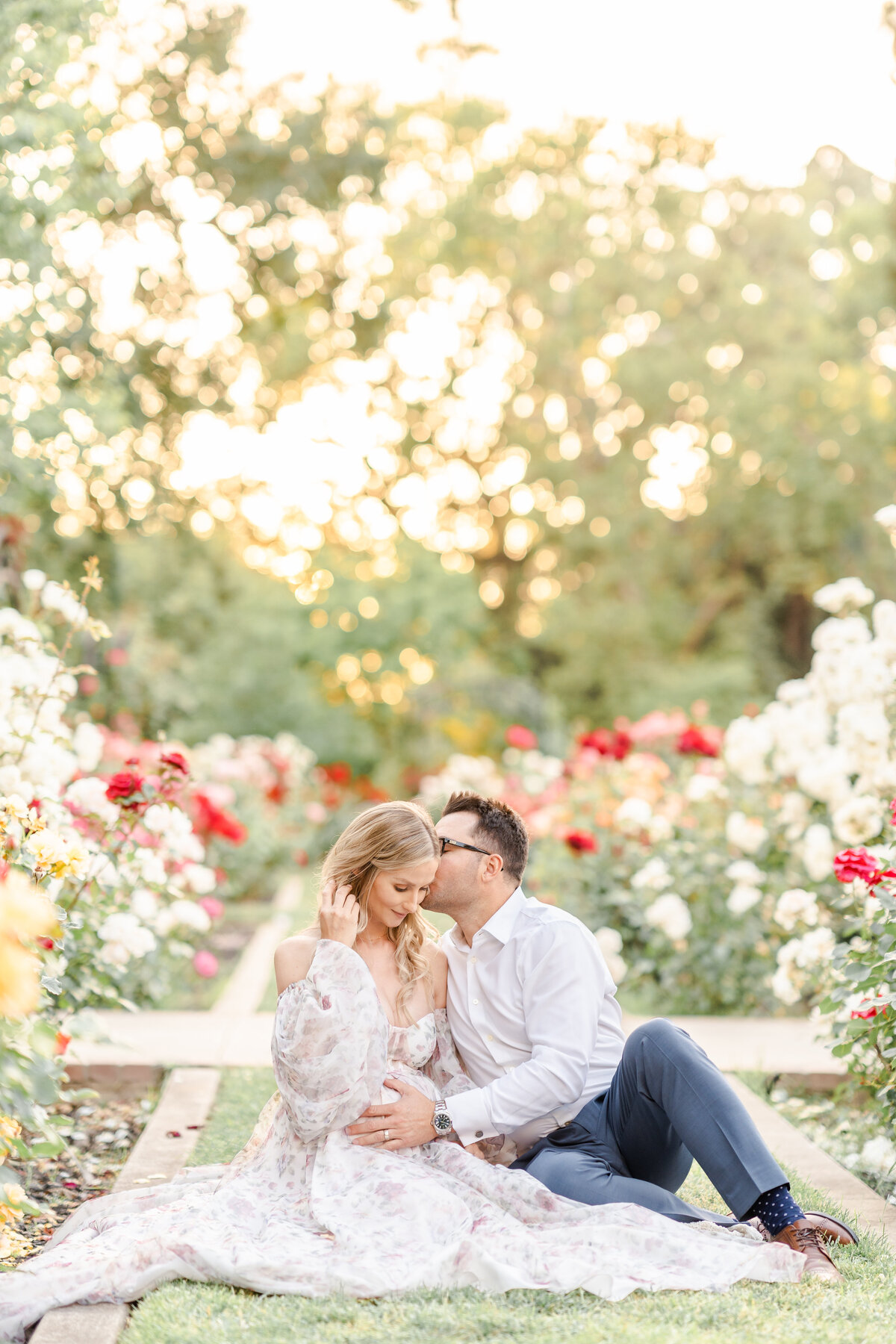 An expecting couple sits together in a rose garden while the man whispers gently into the woman's ear and holds her beautiful baby bump photographed by Bay area photographer, Light Livin Photography.