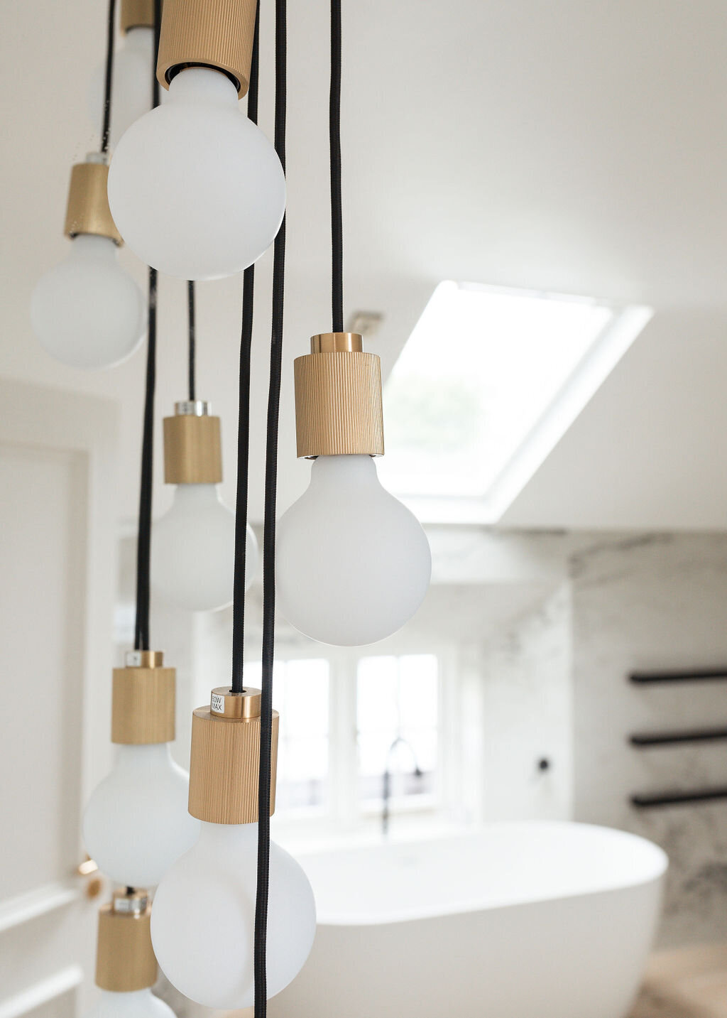 Modern light fixtures with opaque bulbs and brass fittings hang from the ceiling in front of a large skylight window. In the background, a freestanding bathtub sits in a bright, minimalist bathroom.