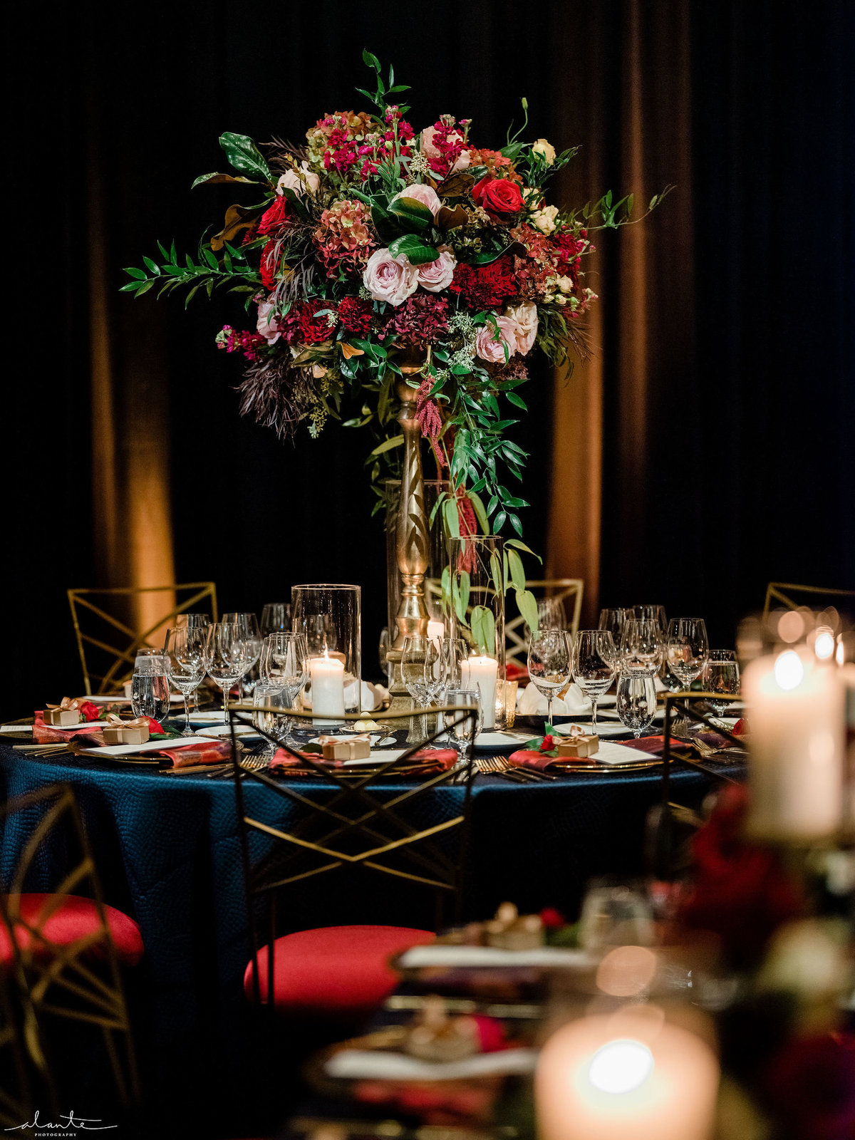 elevated winter flower centerpiece in deep red on table with navy blue linen, red seated chairs, blue draping